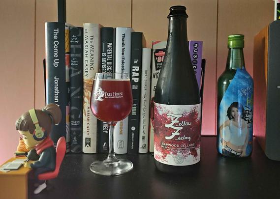 A 5oz stemmed glass filled with beer and a 500ml bottle sit on a shelf. The glass has the Sapwood Cellars logo (half hop/half acorn) w/ antlers on top. Behind on the shelf are various books on music and music artists, and a soju bottle featuring Hyori. A LoFi Girl figure is to the left.