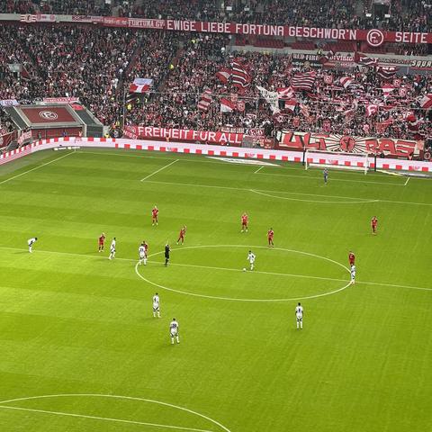 In der Düsseldorf Arena trifft der HSV auf die Fortuna Düsseldorf. Man sieht die Spieler im Vordergrund und im Hintergrund die Düsseldorfer Südkurve.