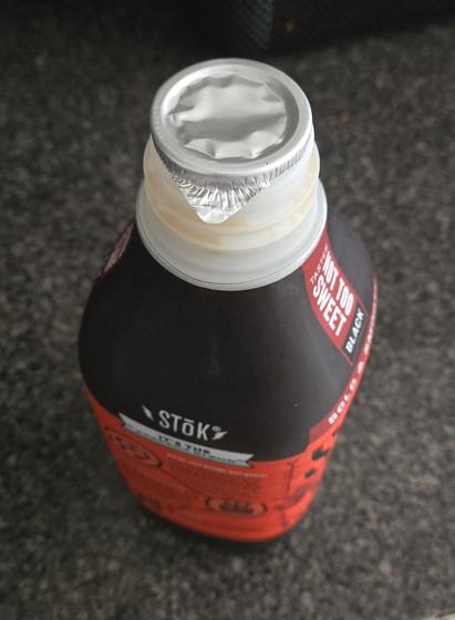 A plastic bottle of cold brew coffee (Stok brand, "not too sweet") is sitting on a mottled gray kitchen counter. The lid has been removed, revealing a metal safety seal over the mouth of the bottle. The seal has a small pull tab on one side.