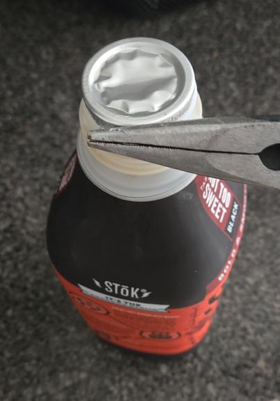The same bottle of cold brew coffee, now with a pair of needle-nose pliers clamped over the pull tab on the metal seal, perpendicular to the tab so that the maximum surface area of the pliers' teeth is on the tab.
