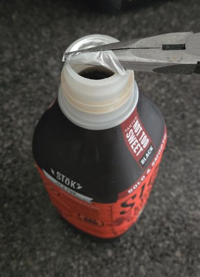 The bottle of cold brew coffee, but this time the needle-nose pliers have been turned while still gripping the pull tab so that the metal seal is wrapping around them and has been pulled open, revealing the delicious beverage beneath.