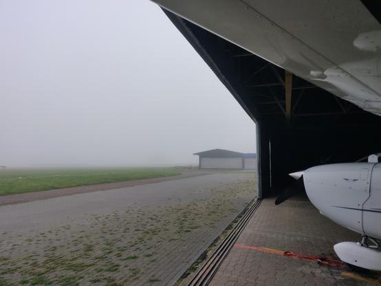 The wing and nose of a light high-wing aircraft sitting in the hangar with the doors open. Fog and a few nearby building visible outside.