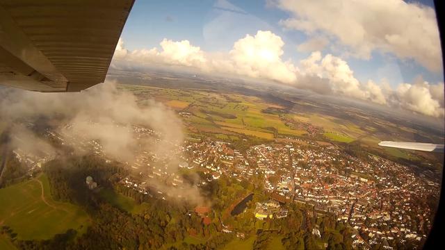 View from the right window of a high-wing light aircraft, small puffy clouds are low above a small town, with a castle and a building with a green dome, fields and more cumulus clouds under a blue sky in the background.