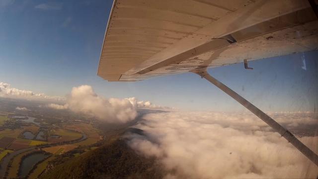 View from the left window of a high-wing light aircraft, blue sky, dense low clouds are bunched up from the right against a mountain ridge. A river and some lakes are sunlit on the left side of the ridge.