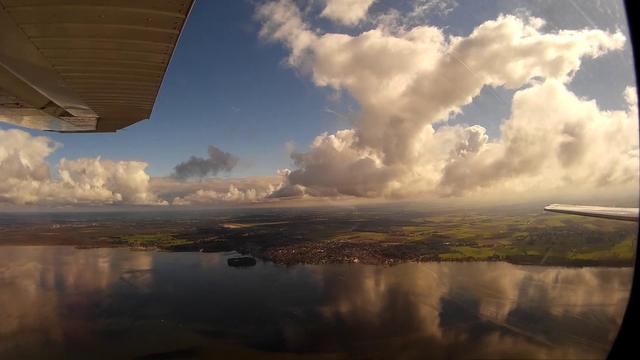 View from the right window of a high-wing light aircraft: blue sky with lots of dramatic cumulus clouds is reflected in a lake. A small coastal town, a military airfield and a small island, connected to the mainland with a bridge, are visible.