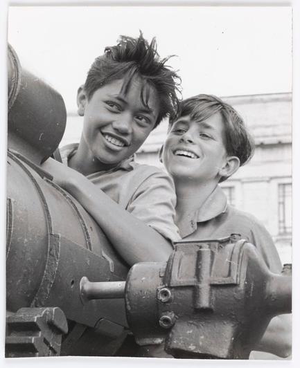 The image captures a candid moment between two young boys, exuding joy and camaraderie. They are standing beside an old-fashioned machine with visible mechanical components like bolts and levers, which gives the photo a sense of nostalgia or historical context. The boy on the left is playfully leaning against the machinery, his hair tousled by wind or perhaps playful movement. His smile is wide and genuine as he looks directly at the camera, radiating warmth.

The other boy stands to one side with an equally cheerful expression, looking in a direction not visible to us but clearly engaging with something amusing. Their clothing suggests they might be on a break from playing outside; both are dressed casually yet neatly for outdoor play or work.

In the background is a blurred image of what appears to be classical architecture, possibly indicating that this scene takes place near a historical site or within an urban environment where such structures coexist with more modern elements. The black and white tone adds a timeless quality to the photograph, emphasizing textures like the boys' clothing fabric and machinery's metal surfaces.

Overall, this image evokes feelings of innocence, friendship, and the simple pleasures found in everyday life during what could be mid-20th-century New Zealand.