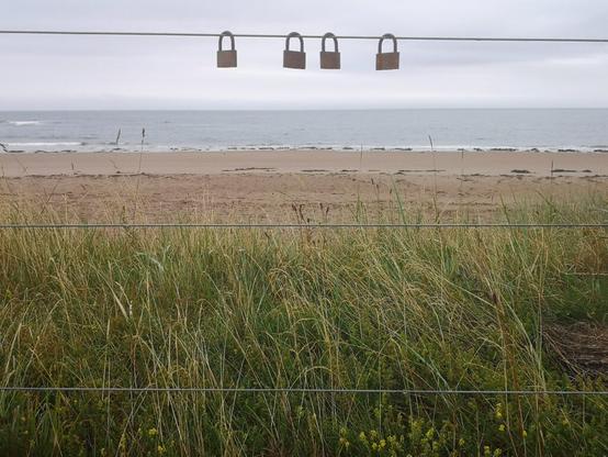 An einem Stahlseil am Strand, hängen vier Vorhängeschlösser, von anderen menschen die schon hier waren. Auf diesen sind verschiedene Sachen eingraviert