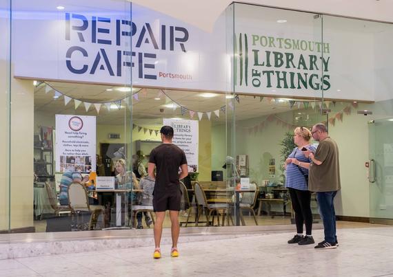 The glass shopfront of Share Portsmouth allows passers by to look and see the Portsmouth Library of things and repair cafe activity going on inside. There is bunting, chairs and tables and people borrowing and getting items repaired inside.