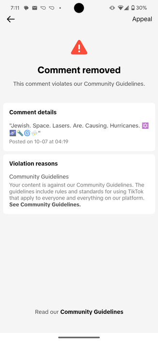 A TikTok notification showing that a comment has been removed for violating community guidelines. The comment details state: "Jewish. Space. Lasers. Are. Causing. Hurricanes." with various emojis (space, magnifying glass, swirling wind, rain cloud, and the Star of David). The post was made on 10-07 at 04:19. The reason provided is that the content goes against the community guidelines, which apply to all users and content on the platform. There is an option to appeal the removal at the top of the notification.