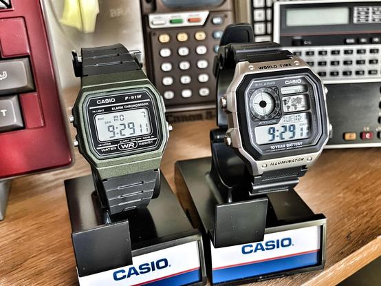 A close-up shot of two iconic digital watches side by side on a clean surface. On the left is the Casio F91W, a simple and classic black digital watch with a slim profile, known for its durability and minimalist design. On the right is the Casio 'Royale' AE-1200, a larger, more feature-rich digital watch with a silver-colored bezel and world time function. The 'Royale' features a prominent screen with multiple time zone displays, a map, and a robust build, while the F91W has a straightforward time display and minimalistic button layout. Both watches are unpretentious yet reliable pieces of everyday tech.