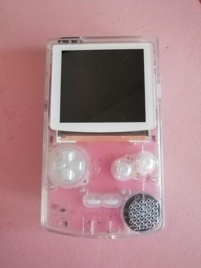 photo of a Gameboy colour hardware replica made by FunnyPlaying. It's on a pink desktop mat and it has a pink pcb which you can see through its white transparent shell. The screen has a white border and the buttons have a transparent pearl shine