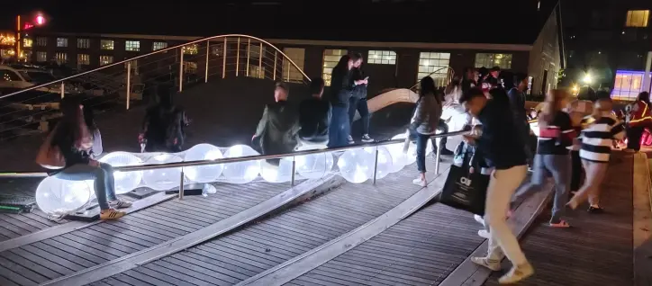 A picture of a string of large lit glowing balls and people are sitting on them. It's on a wooden deck shaped like a wave.