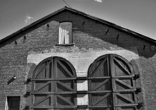 Old, historical brick building for trains. Peaked roof. Double arched wood & iron barn-like doors - where trains can go into in times gone past.
Black and white photo.