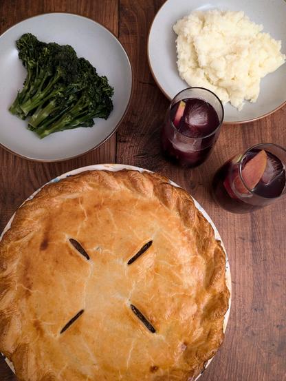 A rectangular photo of some food and drinks on a brown wooden background. There is a golden-crusted pie, a bowl with broccoli, a bowl with mashed potato, and two high-ball glasses filled with a red wine-coloured liquid, apple and orange slices, and ice.