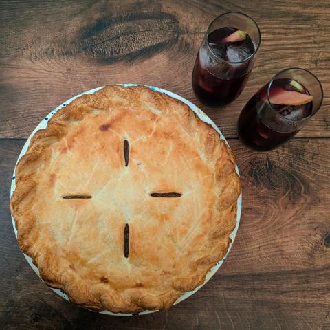 A square photo of a golden-crusted pie and two high-ball glasses filled with red wine, apple and orange slices and ice, sitting on a brown wooden background.