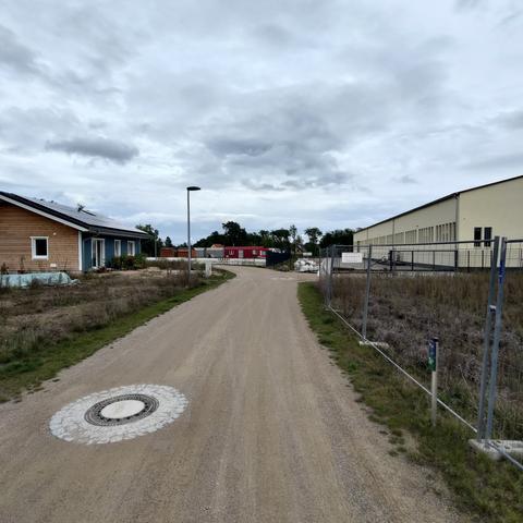 In the middle of a dirt road and surrounded by a wide ring of cobblestones is a rather new manhole cover. To the left of the road is a wooden house, to the right a large hall behind an empty building plot with a construction fence. A cloudy sky is overhead.
