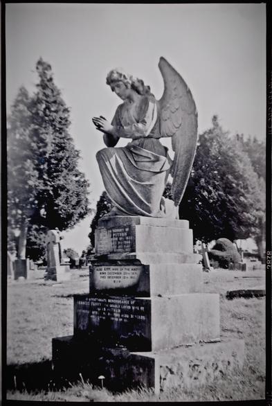 A grainy black and white image of a family gravestone in the shape of a praying angel.