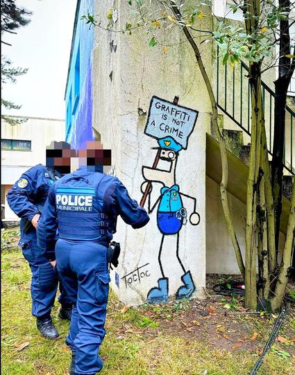 Streetart. A funny mural with a cartoon-style stick figure was sprayed/painted on a gray street wall. He has a protruding lower jaw, one large and one small eye, circles for hands and wears a blue uniform with shorts, a police cap and handcuffs on his belt. He is excitedly holding up a sign that reads: "Graffiti is not a crime". In the photo, two real police officers in blue uniforms stand in front of the wall and inspect the artwork. (The mural was commissioned for a street art festival and is therefore "legal")
Info: The anonymous artist "TocToc" is a French freelance illustrator and video artist. He exhibits his work in galleries around the world. In 2012, he created the simple figures DUDUSS when he was an art student and still sprays it on the walls of cities today.