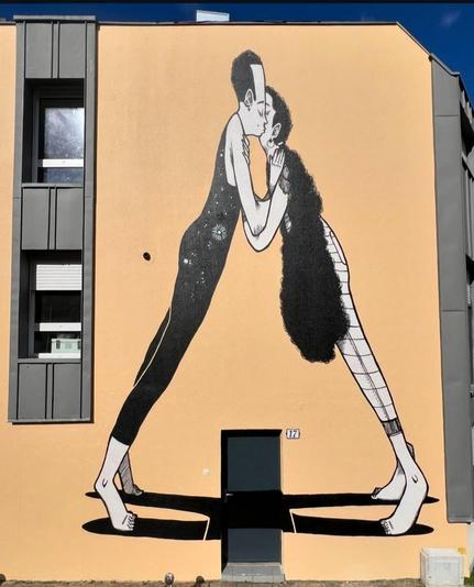 Streetartwall. On the outside wall of a two-story modern building (with a row of windows on the left side), a wonderful mural with two large kissing figures has been sprayed/painted. The background is orange and the figures are designed in an incomparable reduced, comic style in black, gray and white. The figure on the right is a girl with long, curly, black hair that falls around her body in a long braid, almost covering it. She is wearing tight trousers. Opposite her is a man in a tight, long black bodysuit, with black hair and a high forehead. They are kissing and touching each other with their arms, but they are standing on tiptoe far apart so that their bodies look like a triangle. Their shadows cross over the (real) dark entrance door. A wonderfully ingenious and loving mural.
Info: Alex Senna is a famous Brazilian street artist who creates vivid expressions of human love, romance, and relationships through youthful black and white illustrations.