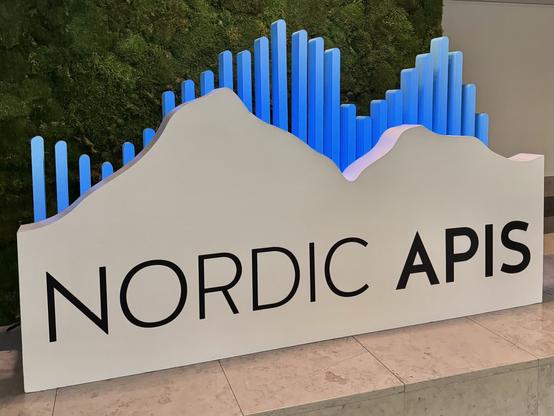 A sign displaying the text Nordic APIs with a stylized mountain design in blue and white, set against a green moss backdrop.