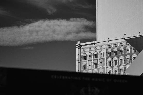 The black and white image shows a partial view of a building facade, possibly printed on a large banner or billboard. It features a classic architectural style with rectangular windows and decorative elements. The upper part of the image shows a cloudy sky, and there is a dark, out-of-focus area at the bottom, which appears to have some text but is mostly obscured.