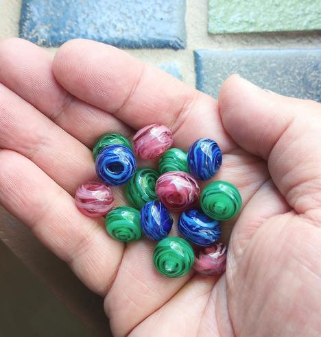 Handmade lampwork glass beads in transparent blue, green and dark rose, with added strands of opaque white, in the palm of my hand.