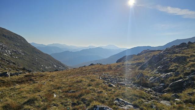 Mountain landscape with grassy slopes and rugged rocks. No path, nor human or animals are visible. Hazy blue peaks are visible in the distance under a clear sky. Sunlight brightens the scenic view, highlighting the rugged terrain and serene atmosphere.