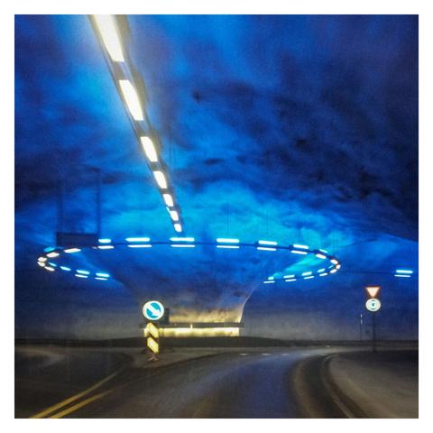 Square color image with a white frame showing a roundabout deep in the mountains of Norway. The image is somewhat blurry, but the road, roundabout, lighting, and traffic signs are still clearly visible. The photo was taken from a moving car. The roundabout is ahead and has three exits, one to the right and one to the left. From the roundabout, a kind of pillar extends into the ceiling, becoming significantly wider (about double the diameter) as it goes up. A yellow light ring is attached to the lower end, illuminating the lower section of the pillar. Around the entire ceiling of the roundabout, blue (upward) and white (downward-facing) lights are installed, casting a blue glow with many dark areas across the ceiling. A row of lights extends from the top left corner of the image directly toward the center of the roundabout, following the road. Two traffic signs are visible in front of the roundabout: a blue sign with a right arrow directly in front of the roundabout, and a yield sign on the right side with a blue roundabout sign.