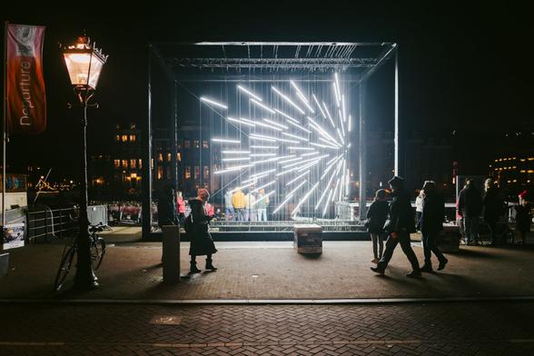A box contains beams of light that look like a firework mid-explosion. People walk past and look at it.