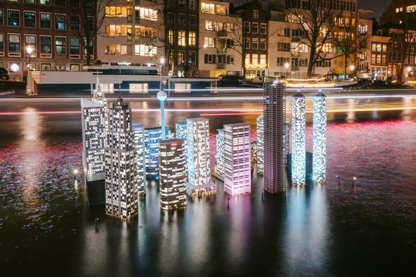 Lit up models of buildings sit in a canal in Amsterdam. Barges pass by in the background leaving light trails.