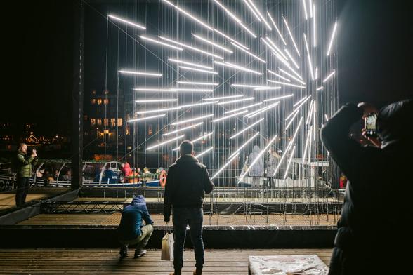Close in on a light installation showing beams of light in a box mimicing a firework mid-explosion. People take photos.