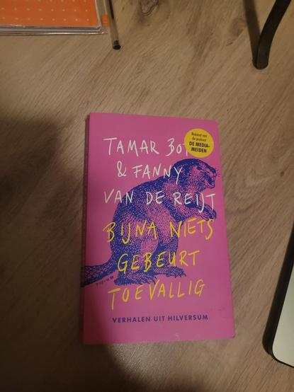 Picture on a wooden floor of a Dutch book cover (pink with a blue lithografie of an animal) "tamar boy and fanny van de reijt (authors), bijna niets gebeurt toevallig (title)", by the editions verhalen uit Hilversum