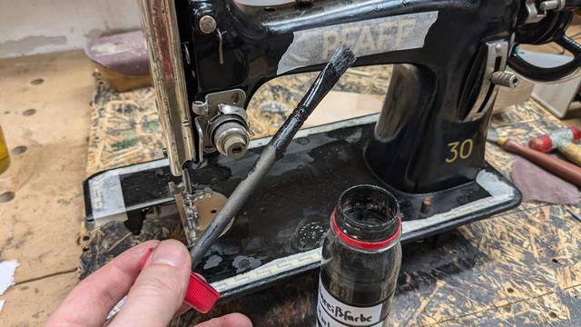 The machine is standing on an OSB workbench top, and all its golden decals have been taped off with masking tape. A plastic bottle with black shellac stands in front of it, and a hand holds a small paint brush with black paint into the frame.