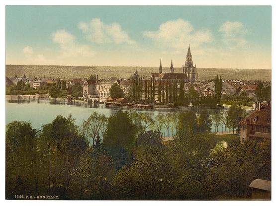 The image depicts a picturesque townscape, possibly from the late 19th or early 20th century based on its style. It showcases a serene lakeside town with various buildings in different architectural styles, including what appears to be historic churches and more modern structures alongside trees that line the shoreline of Lake Constance (Bodensee). The sky is partly cloudy, allowing for sunlight to illuminate parts of the scene. Notable features include tall spires of a church or cathedral dominating the skyline with its distinct towers reaching towards the heavens. A river or lake stretches across much of the landscape, reflecting light blue hues typical of water bodies in such prints from that era. The vegetation is lush and green, suggesting it might be late spring to early summer. There's no direct human activity visible, which adds a timeless quality to the image. It appears as if this could have been captured by P.Z., Konstanz based on the text at the bottom of the print indicating its historical context or origin.