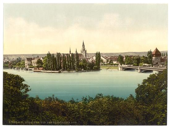 The image showcases a picturesque view of Constance, located in Baden, Germany. It appears to be taken from the Seestrasse area and captures an expansive panorama overlooking Lake Constance (Bodensee). The lake is prominently featured with its turquoise waters stretching across the center of the composition.

Various architectural elements enhance the historical ambiance of the scene: a prominent Gothic church or cathedral stands out in the middle distance, accompanied by other spired buildings that suggest medieval and Renaissance influences. Greenery from trees lines up along the waterfront's edge, adding to the image’s charm.

In the foreground, dense foliage frames the view, possibly indicating a vantage point like an elevated hillside overlooking this serene town and lake landscape. A bridge spans across one side of the water body, hinting at connectivity between different parts of Constance. The overall color palette is dominated by natural greens from vegetation, earthy tones from buildings, and blue hues from the lake.

The vintage aesthetic suggests that it could be a photograph or postcard dating back to approximately 1890-1900 as per the caption 'From the Seestrasse, Constance (i.e. Konstanz), Baden, Germany between ca. 1890 and ca. 1900'. The image provides a glimpse into early photographic techniques used to capture such landscapes with great detail and color [...]