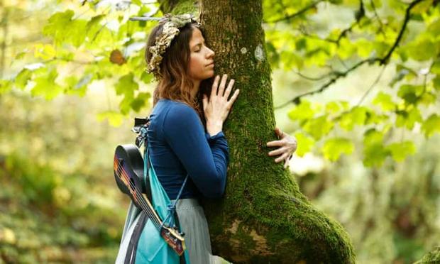 A woman wearing flowing clothing in various shades of blue, a garland on her head, and a ukulele slung across her back, hugs a moss-covered tree in woodland.