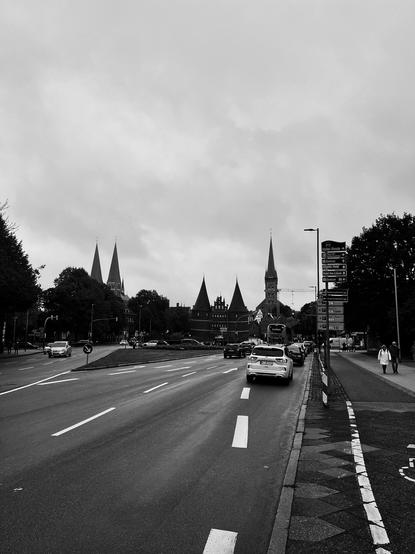 A black and white image of a street with traffic, featuring notable church spires and historical buildings in the background. The sky is overcast, and there are pedestrians on the sidewalk. Directional signs are visible on the right.