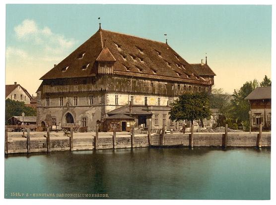This image depicts a historical building situated by the water, likely in Constance (Konstanz), Baden, Germany. The architecture is characteristic of German medieval buildings with its steeply pitched wooden roof and dark timber framing. Several windows are visible on multiple levels of the structure, suggesting it may have been used for residential or administrative purposes.

A body of calm blue-green water stretches out in front of the building, possibly a canal or part of Lake Constance, which is considered one of Europe's largest lake systems. To the right and left sides of the image are more modern buildings with less intricate design work compared to the central structure. A sign on the facade indicates it might have had some significance as "Konstanz Das Gounciliumgebäude," likely translating to "Constance The Council Building."

The photo appears aged, possibly dating from around 1890 to 1900 based on its condition and style of printing. It is labeled with the number "1548" in a red stamp at the bottom right corner, which might be an internal cataloging or identification mark.

In summary, this image captures a historical moment where traditional German architecture meets calm waterside living in what was likely an important community space for local governance during that period.