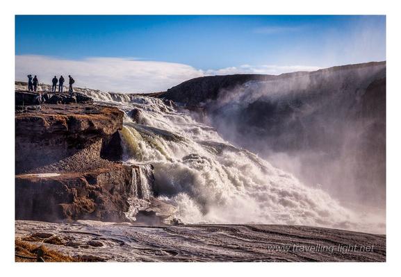 Side view photo of a large waterfall cascading down a stepped slope, with people standing beside the top of the fall, against the sky.
