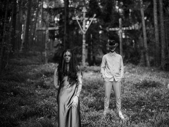 The image is a black-and-white portrait of a couple in a forest. In the foreground stands a woman with long hair, tattooed arms, and a veil partially covering her face. She wears a simple, dark dress and looks directly at the camera. In the background, slightly out of focus, stands a man wearing a hat and a shirt, with his face obscured. The atmosphere is mystical and dark, and the setting appears remote and tranquil, with several wooden crosses in the background, adding a symbolic and slightly eerie character to the scene.