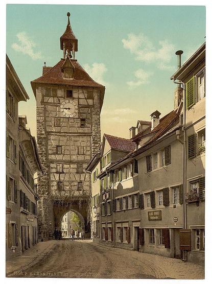 The image depicts an old, picturesque street in Constance (now Konstanz), Germany. The central feature is the Schnetzthor, a historic city gate with a prominent clock tower rising above it. Constructed from stone blocks, the structure exhibits traditional architectural elements such as timber framing and decorative stonework.

Surrounding buildings are multi-storied, exhibiting classic European design with shuttered windows and pitched roofs, indicative of early to mid-19th-century architecture. The street itself is cobblestone, curving gently around the tower's base, leading towards an open area where a few figures can be seen in the distance.

The photograph has been colored sepia-toned, giving it a vintage feel consistent with its estimated date between 1890 and 1900. This coloration suggests that the image may have undergone post-processing to enhance or recreate the historical ambiance of the scene.

Additional context indicates that this print is part of a series showcasing views from Germany during the late 19th century, as identified by "The Schnetzthor" being listed alongside another photograph with similar dating. The overall composition and preservation quality suggest it might be used for educational or research purposes to understand historical architecture and urban planning in German cities at that time period.

Please note that any specific modern-day loca [...]