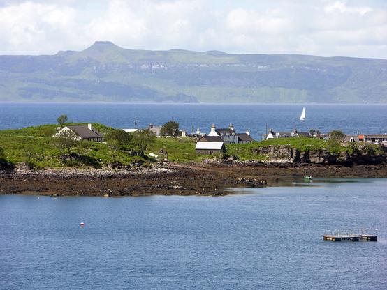 Ard-dhubh. The image shows a narrow promontory of land stretching across the centre of the frame with sea this side of it and beyond it. The tide is out, leaving a broad margin of brown seaweed on view. The village comprises a line of cottages and houses on the promontory, partly concealed by rises in the landscape. In the distance is land rising to hills including an angled summit. The scene is in sunlight.