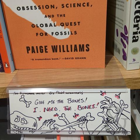 The Dinosaur Artist: Obsession, Science, and the Global Quest for Fossils by Paige Williams with its orange jacket is on display at Barnes & Noble. Beneath the book, attached to the shelf, is a staff recommendation plaque with a drawing. The drawing has the text "GIVE ME THE BONES! I  . NEED . THE . BONES!" There is a drawing of a t-rex skull on the dar left and to the right four hands gripping or trying to grip bones.