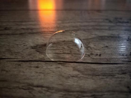 Eine Seifenblase "sitzt" auf einem Holztisch. Im Hintergrund ist das warme Licht einer Kerze zu sehen. ~~~ A soap bubble "sits" on a wooden table. The warm light of a candle can be seen in the background.