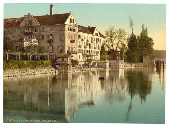 This image depicts a picturesque scene of the Konstanz Hotel on an island, located in Constance (now known as Konstanz), Baden, Germany. The architecture showcases traditional European design with its ornate facades and multiple levels featuring balconies and windows. A serene body of water reflects the building's facade and surrounding trees, enhancing the tranquility of the setting. In the background, a smoking chimney suggests industrial activity or heating from an older time period.

The photo appears to be vintage, indicated by its coloration typical of early photographic processes like albumen silver prints with hand-tinted colors. The inclusion of "Hotel" on the building's facade makes it clear that this structure serves as lodging for visitors. Additionally, the date stamp at the bottom reads 1547 P.Z., which could be referencing a specific catalog or archive number.

This postcard-like image captures not only an architectural and natural beauty but also provides insight into tourism practices of its time by highlighting local attractions such as this hotel island in Lake Constance.