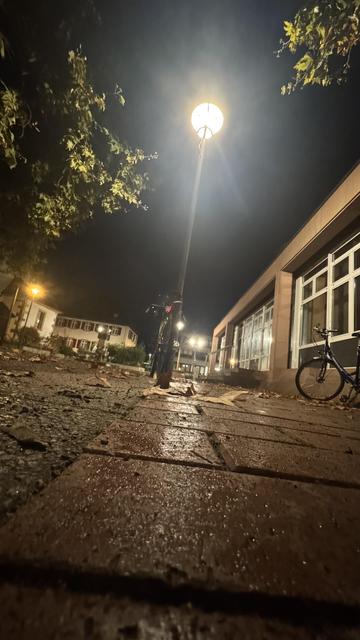 Eine wenig beleuchtete Nachtszene mit einem Bürgersteig mit nassen Blättern und Backsteinpflaster. Ein Laternenpfahl beleuchtet das Gebiet, und zwei Fahrräder sind in der Nähe aufgestellt, mit einem Gebäude im Hintergrund.