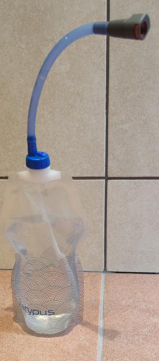 A Platypus soft bottle which is about 90% full of water stands upright. The standard screw top has been replaced with a modified Source top and a hydration pipe.
