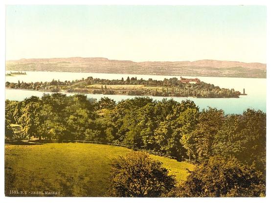 The image depicts a picturesque view of an island situated in the middle of a large body of water, which appears to be Lake Constance given its geographical context. The vantage point is from an elevated landmass with greenery and trees dominating the foreground.

In the distance on the right side of the lake, there's a prominent building complex set amidst verdant landscapes, suggesting it could be a manor or castle-like structure. This central island is connected to the mainland by bridges or causeways, indicating accessibility for visitors.

The sky above has a warm tone, possibly due to the coloration typical of vintage photographs from around 1890-1900. The overall impression is one of tranquility and natural beauty, with human habitation harmoniously integrated into nature's landscape.
