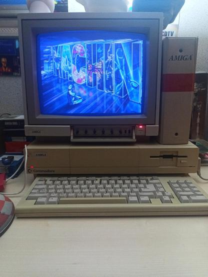 Un moniteur 1081 posé sur un Amiga 1000, avec un clavier en premier plan et un classeur "Amiga" posé à côté du moniteur. Le moniteur affiche une jeune fille assise en tailleur devant un logo "MACHINE" écrit très gros, le tout avec des teintes bleutées.