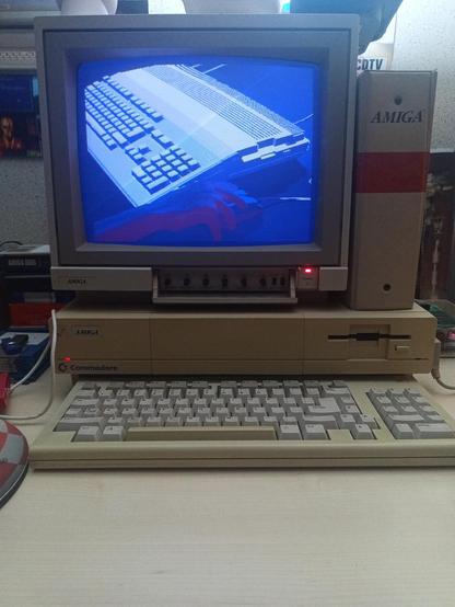 Un moniteur 1081 posé sur un Amiga 1000, avec un clavier en premier plan et un classeur "Amiga" posé à côté du moniteur. Le moniteur affiche un clavier d'Amiga 500 et une main qui tient une souris, le tout avec des teintes bleutées. On dirait presque une photo.